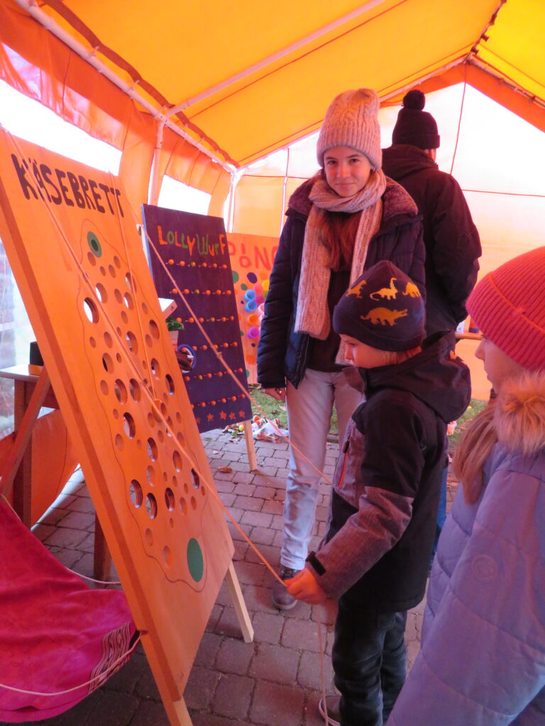 Ein Kind und eine jugendliche Betreuerin an einem Geschicklichkeitsspiel in einem großen orangen Zelt