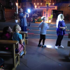 Pfarrer Pauly leuchtet mit einer Taschenlampe durch die dunkle Kirche, die Kinder schauen dem Lichtstrahl hinterher